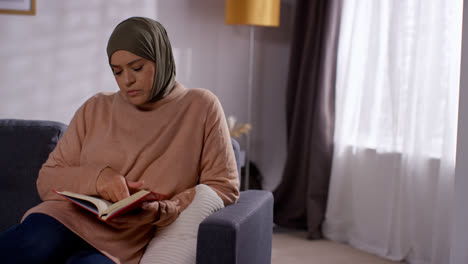 Mujer-Musulmana-Con-Hijab-Sentada-En-Un-Sofá-En-Casa-Leyendo-O-Estudiando-El-Corán-2